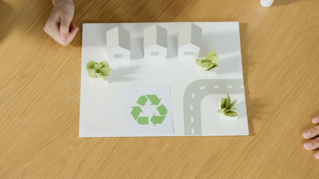 Modello di città sostenibile con case in miniatura e piante, evidenziando il simbolo del riciclo su un piano di lavoro in legno.