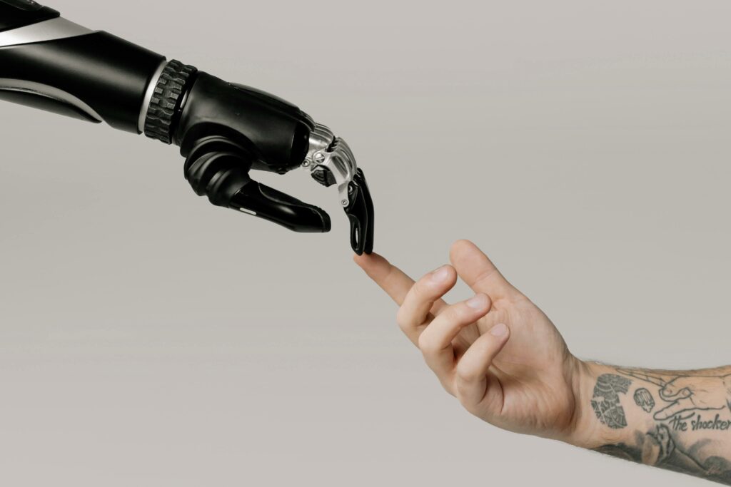 Mano robotica nera e mano umana si sfiorano, simboleggiando l'interazione tra tecnologia e umanità. La mano robotica presenta dettagli metallici e una struttura avanzata, mentre la mano umana mostra tatuaggi, evidenziando la connessione tra il mondo artificiale e quello naturale.