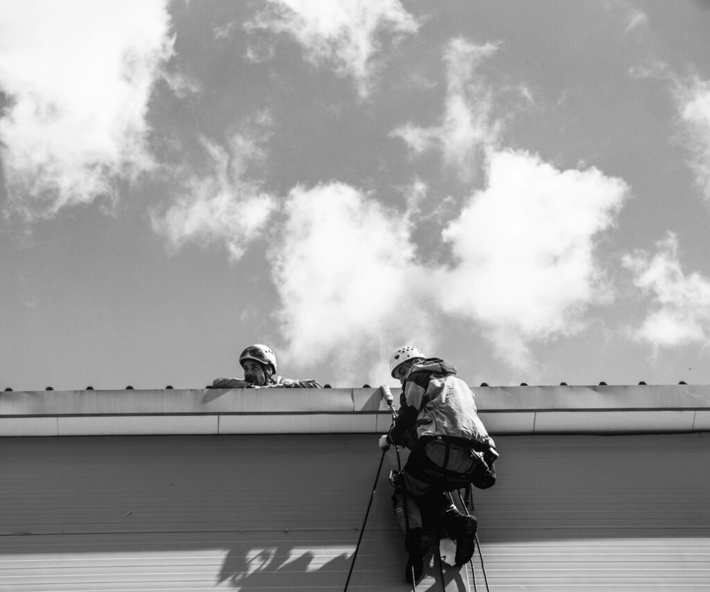 Lavoratori in abbigliamento di sicurezza mentre eseguono operazioni di manutenzione su un tetto, con attrezzatura da arrampicata e caschi protettivi, sotto un cielo nuvoloso.